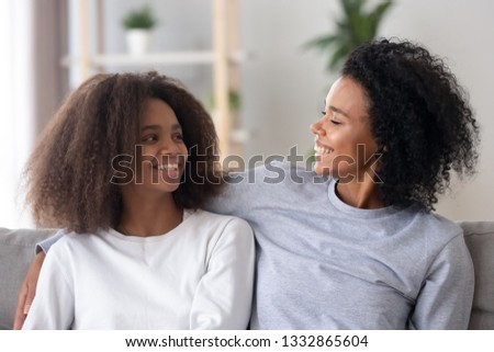 Happy biracial mom looking at teen daughter cuddle her express love, devotion. Older and younger sisters having good relations best friends laughing look at each other sitting on couch in living room Royalty-Free Stock Photo #1332865604