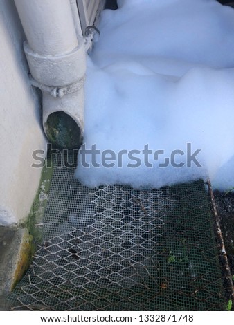 A close up view of white bubbling foam that has come down the main drain pipes and it spilling out at the bottom of the main water pipe and flowing over the metal grids