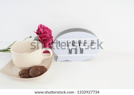 Mother's day.  I love you MOM written at the white oval word board with the carnation flowers and a cup of coffee serve with cookies over the white background