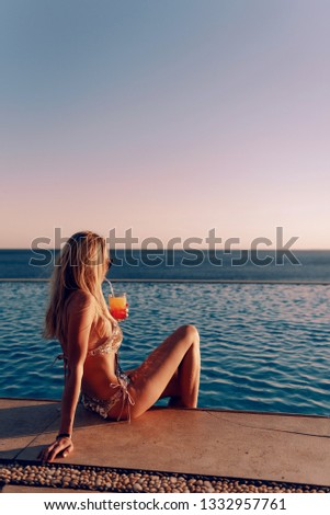 girl in an expensive golden swimsuit with a cocktail in her hands sits at the edge of the pool in the rays of the setting sun