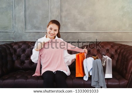 Beautiful Asian woman sitting on the couch.The pink shirt is in his hand.Many shirts And several paper bags on the brown sofa in the room.Woman smiling.Do not focus on the object.


