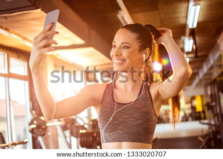 Smiling fitness girl with earphones using smartphone to take a selfie in the gym