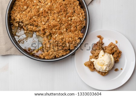 An apple crumble cake topped with a scoop of  vanilla ice cream placed on a white plate next to the baking pan. Side view shot and table top view of the dish.