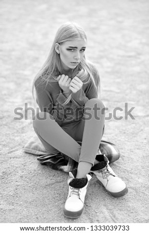 Young blond model with straight hair in bright outfit sitting on street with legs crossed and holding her arms crossed at her chest.