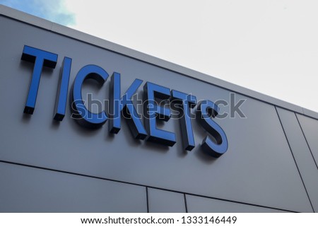 The exterior wall of a modern contemporary metal composite panel commercial building with an illuminated blue lettering tickets sign on the sale booth.  The upward view of an admission office signage.