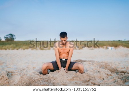 Young topless man sitting on beach beyond sea with legs inside wholes in sand and sculpting figures of sand by hands like child. Hobby, leisure on summer vacation concept. Art person creative activity