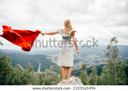 Woman in wild mountains gives distress signal SOS using red cover. Concept of emergency situation during hike in mountains. Barefoot woman stands at stone, waving red blanket and waiting for help.