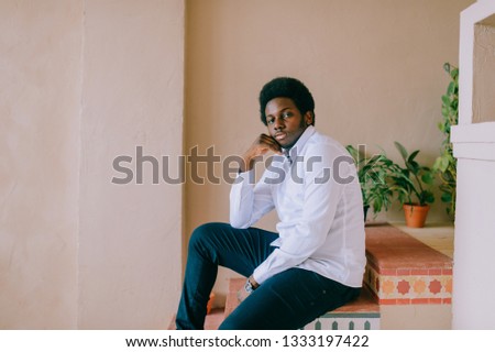 Lifestyle portrait of handsome young african black man sitting in chair  on textured wall with picture on background. Indoor portrait of happy dark skinned nigerian confident man relaxing at home