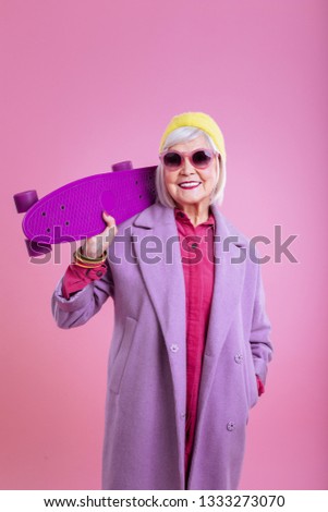 Purple skateboard. Modern retired lady wearing pink sunglasses holding bright purple skateboard