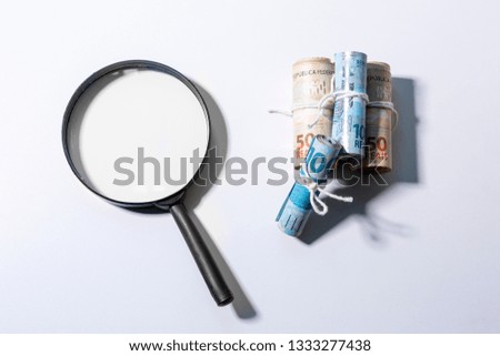 Large magnifying glass next to a stack of rolls of banknotes. Clean studio shot currency with magnify tool against a white background.