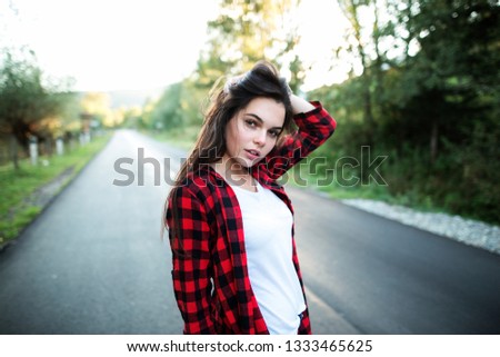 Beautiful happy woman plaid shirt and jeans stand on the road with tree around and looks at the camera