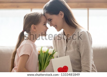 Cute little kid daughter making surprise congratulating happy mom with mothers day birthday 8 march presenting flowers greeting card, smiling loving affectionate family mum and child girl, side view