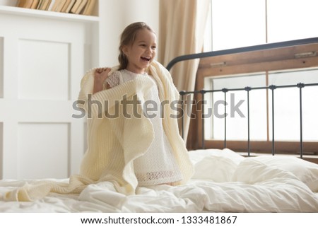 Cute playful happy kid girl covered in knitted plaid laughing having fun sitting on bed, cheerful carefree funny little preschool child feeling joy playing with laughter in bedroom in the morning