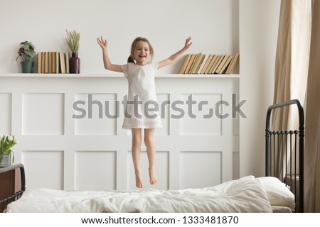 Happy funny little child girl in motion jumping on bed alone flying in air feeling joy, cheerful cute active kid having fun playing laughing in bedroom after waking up, good morning children concept