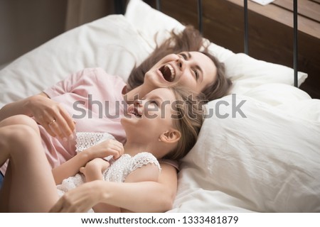 Happy family young mother embracing little kid daughter laughing playing lying on bed, cheerful funny small child girl having fun with smiling mom hugging cuddling enjoying good morning in bedroom