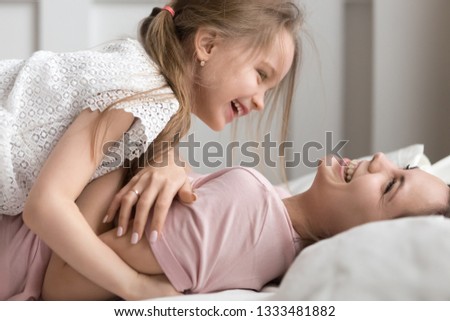 Happy little kid daughter and mother having fun playing with laughter joy tickling in bed, cheerful family funny cute small child girl and mom laughing cuddling together enjoy morning time in bedroom