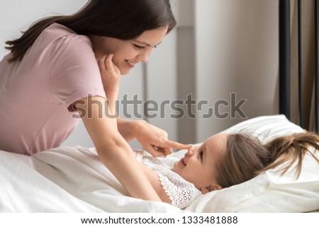 Happy caring mother tenderly waking up cute little kid daughter lying in bed touching her nose, small child girl awaking having fun enjoying family good morning with loving smiling mom in bedroom