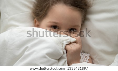 Cute little kid girl looking at camera under blanket lying in comfortable bed on white pillow, pretty child peeking from warm duvet waking up in the morning after sleep cover face top view from above