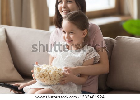 Happy family mom with kid daughter watching tv together with popcorn food sitting on couch, smiling mom and little child girl having fun enjoy television show cartoons movie holding remote control