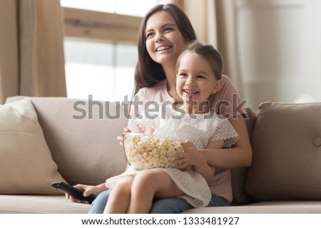 Happy young mom and child girl laughing holding snack popcorn remote control enjoy funny television comedy movie, smiling family mother with kid girl watching fun humor tv show laugh sitting on sofa