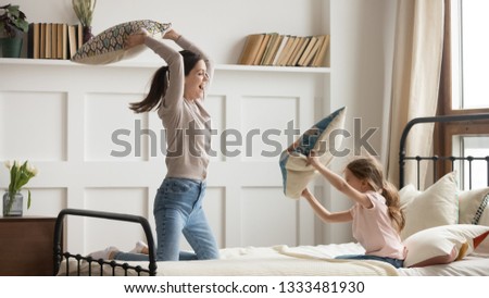 Happy family mom baby sitter and little kid daughter having fun pillow fight on bed, young mother nanny laughing playing funny game with small child girl in bedroom joy leisure morning activity