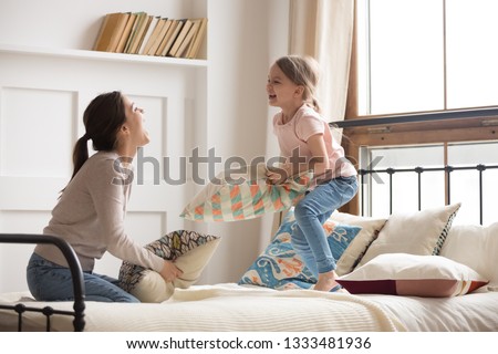 Happy mom elder sister and child girl enjoy funny pillow fight on bed, babysitter mother with little kid daughter having fun together in bedroom, cheerful family play laughing together in the morning