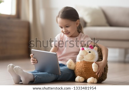 Curious little child girl having fun using digital tablet alone embracing toy sitting on floor, happy preschool smart kid playing with computer looking at screen watching cartoons online at home Royalty-Free Stock Photo #1333481954