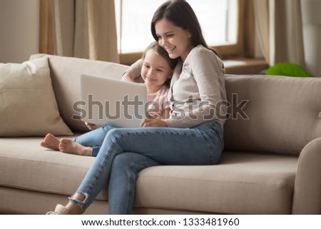 Smiling mom enjoy watching online cartoons making video call on laptop with little child daughter, happy family mother and cute kid girl using computer at home sitting on sofa looking at pc screen