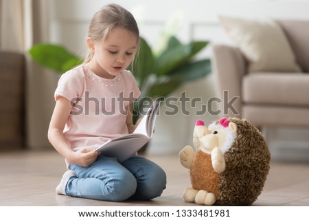 Cute smart little kid girl playing alone reading story to fluffy hedgehog sitting on warm floor at home, funny creative preschool small child holding book teaching toy, children imagination education Royalty-Free Stock Photo #1333481981