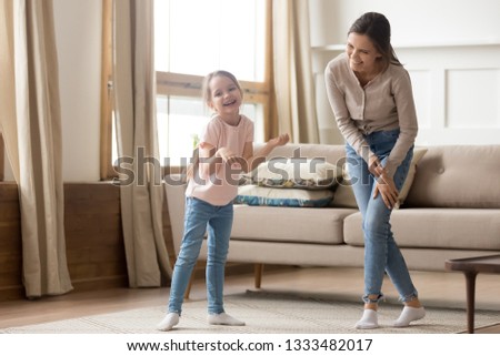 Happy playful cute child daughter laughing enjoy funny home activity with young mom, elder sister or baby sitter, funny little kid girl and cheerful mother having fun dancing together in living room