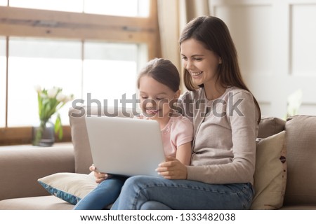Happy family mother and child daughter use laptop computer at home sit on sofa, smiling mom teaching kid girl having fun looking at pc screen doing online shopping watching cartoons making video call
