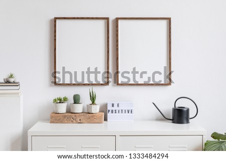 Minimalistic home decor of interior with two brown wooden mock up photo frames , white shelf with books, black watering can, cacti composition, box and home accessories. White walls. Mockup concept.