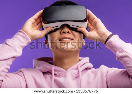Asian young man dressed in white hoodie using virtual reality headset isolated over violet background