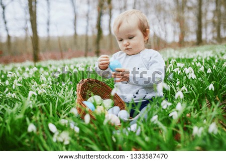 One year old girl playing egg hunt on Easter. Toddler sitting on the grass with many snowdrop flowers and gathering colorful eggs in basket. Little kid celebrating Easter outdoors in park or forest