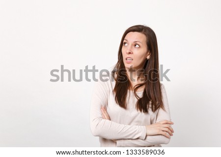 Portrait of puzzled pensive young woman in light clothes looking up, holding hands crossed isolated on white wall background in studio. People sincere emotions, lifestyle concept. Mock up copy space