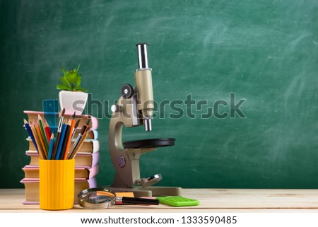 Education and science concept - group of colorful books and microscope on the wooden table in the classroom, blackboard background