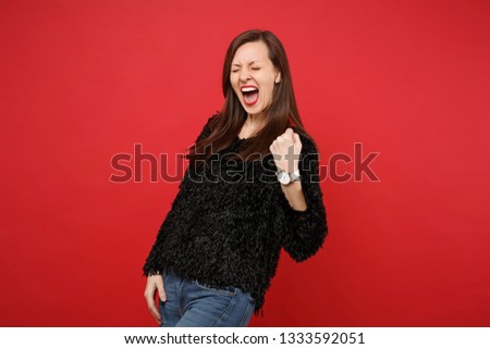Overjoyed young woman in black fur sweater doing winner gesture, keeping eyes closed, screaming isolated on bright red wall background. People sincere emotions, lifestyle concept. Mock up copy space