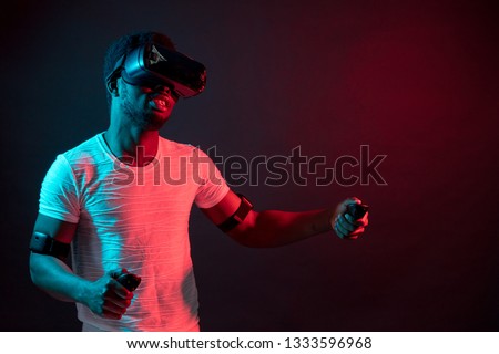 Young man wearing VR glasses watching video or movie, isolated on dark red dual light, testing gadget for virtual reality, isolated on black background with red colour flashes.