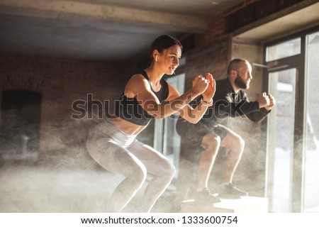 Fit caucasian couple doing squats on boxes in a crossfit style gym, well lit by bright sunlight. Royalty-Free Stock Photo #1333600754