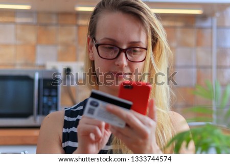 Happy young woman using credit card and phone in kitchen at home