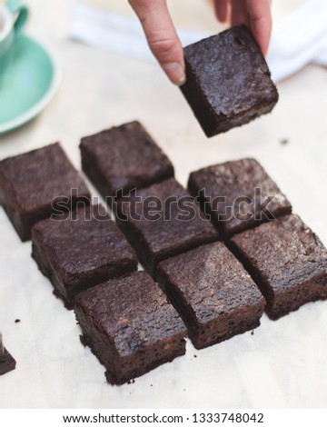 A bright and light picture of a brownie cut into 9 pieces laid on pargament paper, while one piece is being picked up by elegant fingers.