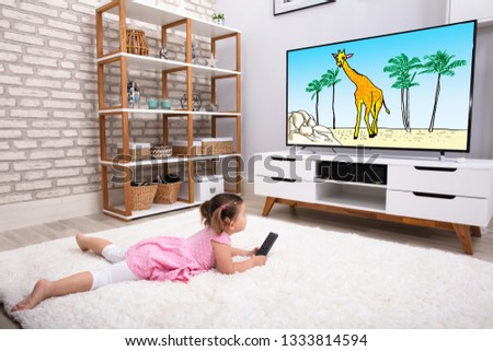 Rear View Of Innocent Girl Lying On Carpet Watching Cartoon On Television