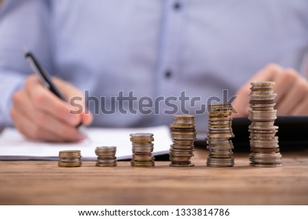 Close-up Of A Businessman Calculating Invoice Using Calculator In Front Of Stacked Coins