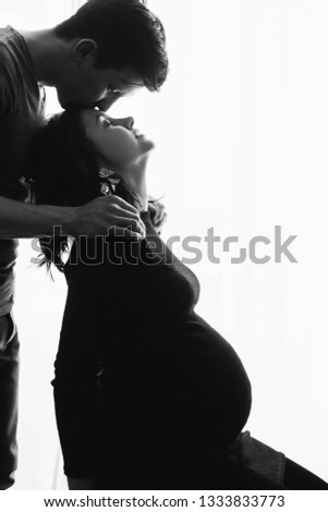 Silhouette of happy husband kissing his pregnant wife in stylish black dress and holding belly bump in light. Fashionable pregnant mom and dad, posing and hugging belly. Parenthood support