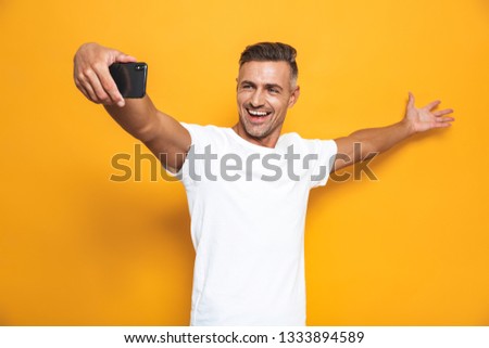 Image of good-looking man 30s in white t-shirt laughing and taking selfie photo on mobile phone isolated over yellow background