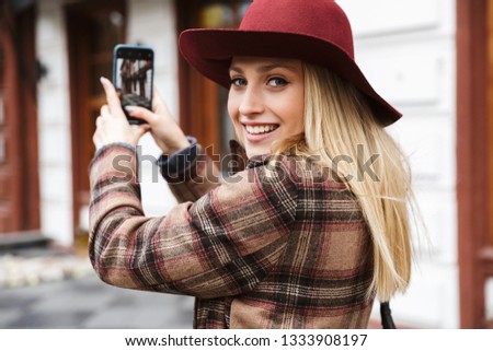 Beautiful young stylish blonde woman wearing a coat walking outdoors, taking photos with mobile phone