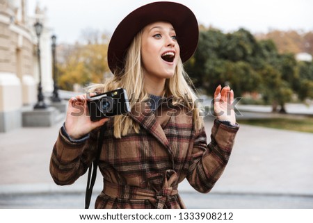 Beautiful young stylish blonde woman wearing a coat walking outdoors, taking pictures with photo camera