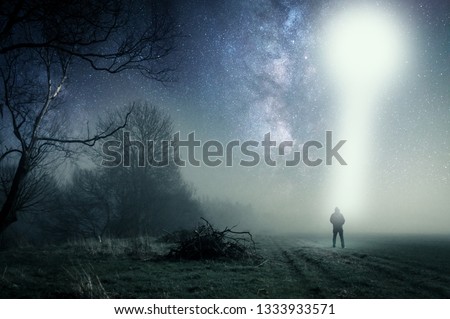 A concept of a UFO above a lone hooded figure with a light beam coming down. Standing on a path on a spooky misty night, with a cold grainy blue edit.