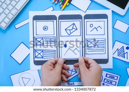 Working desk with hands sorting wireframe screens of mobile responsive website. Developing wireframe sketch layout design mockup on smartphone,tablet screen.