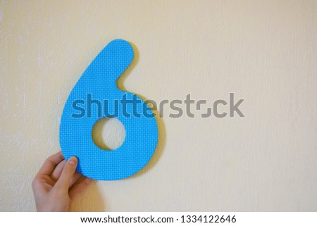 Hand holding blue number six on yellow wall background with copy space for text. 6th anniversary birthday design or educational children toys for learning colors and numbers concept.
